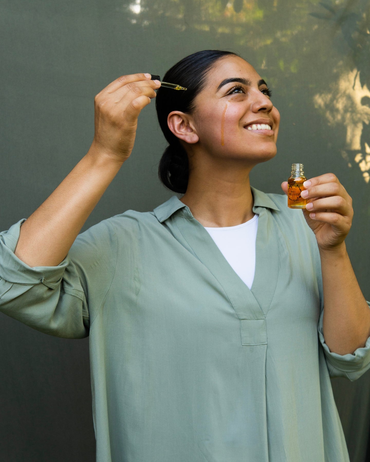 A smiling woman holds the Awake Coffee Serum with a drop of serum running down her cheek.
