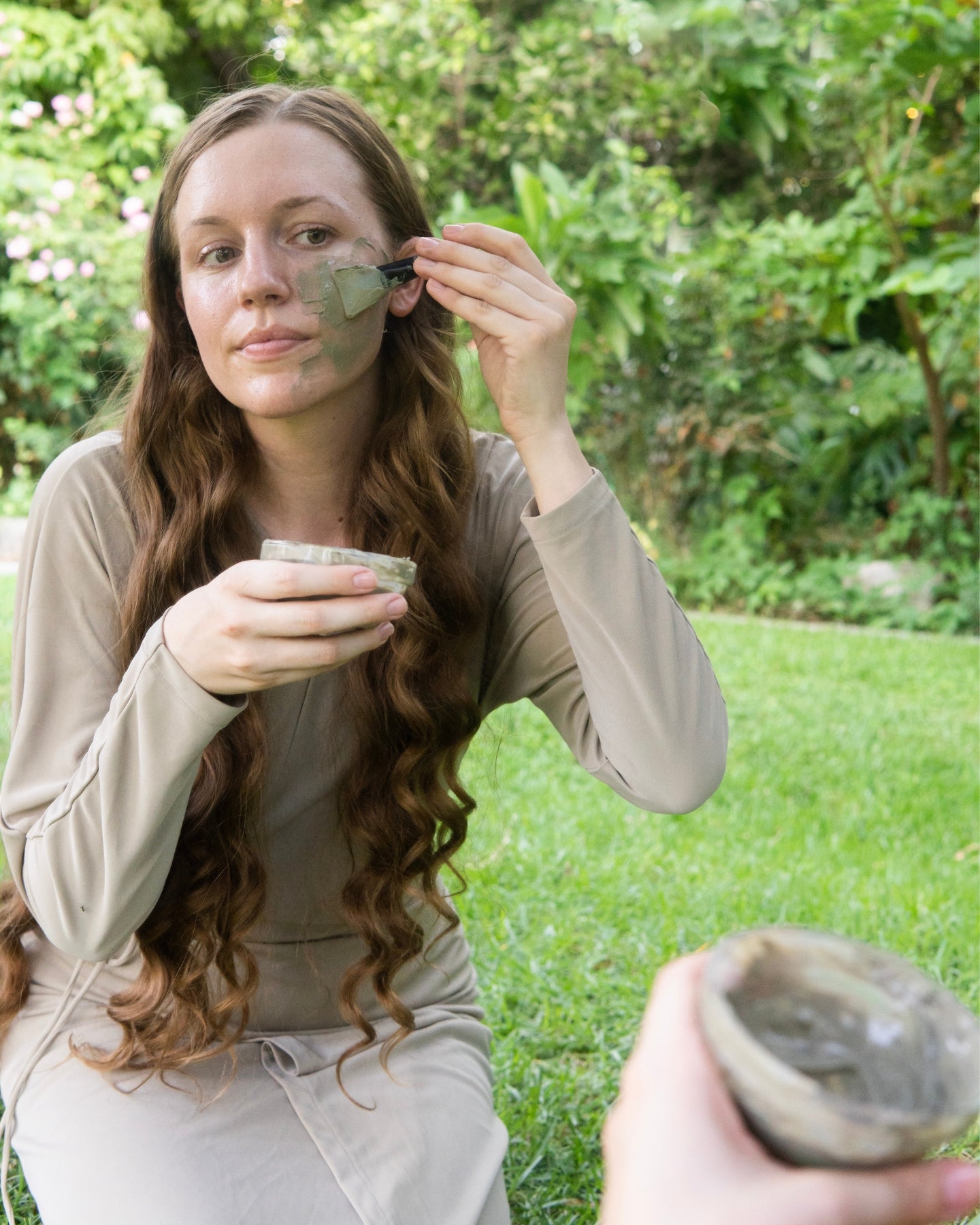 A woman applies the Detox Clay Mask to her face while kneeling in the grass.