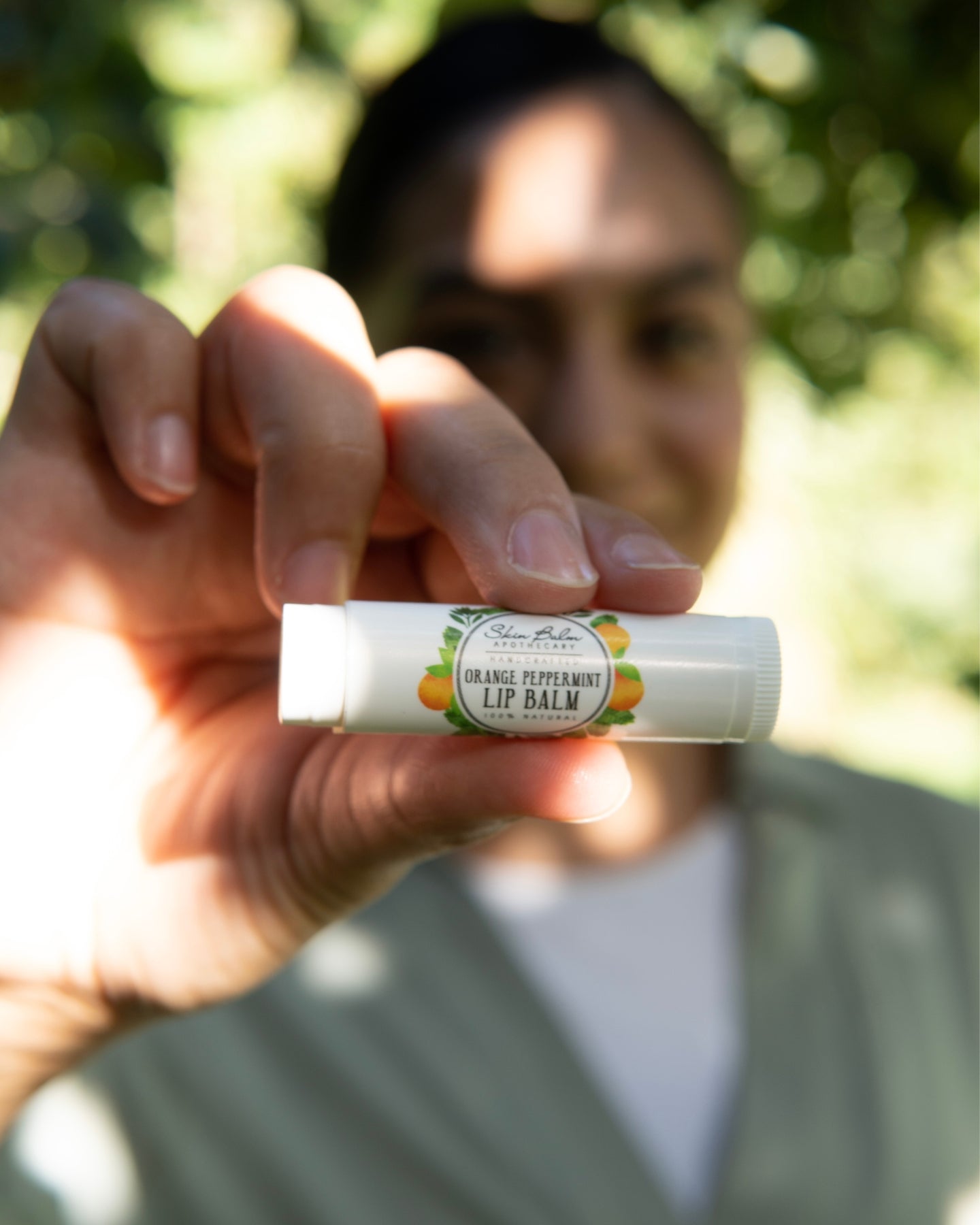 Orange Peppermint Lip Balm focused in the foreground with a woman blurred in the background.