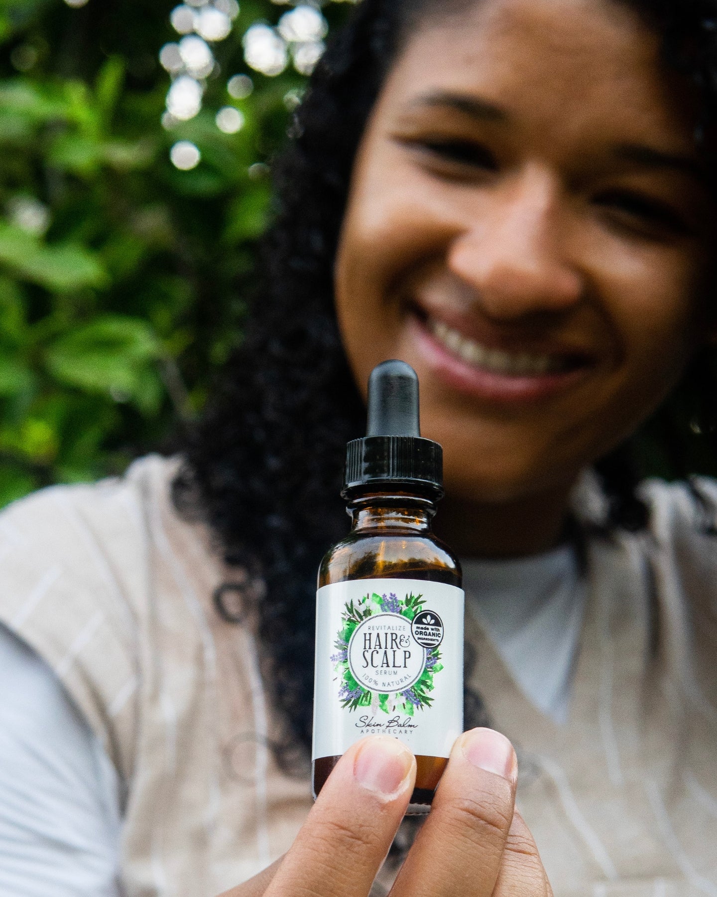 Revitalize Hair & Scalp Serum focused in the foreground with a smiling woman blurred in the background.
