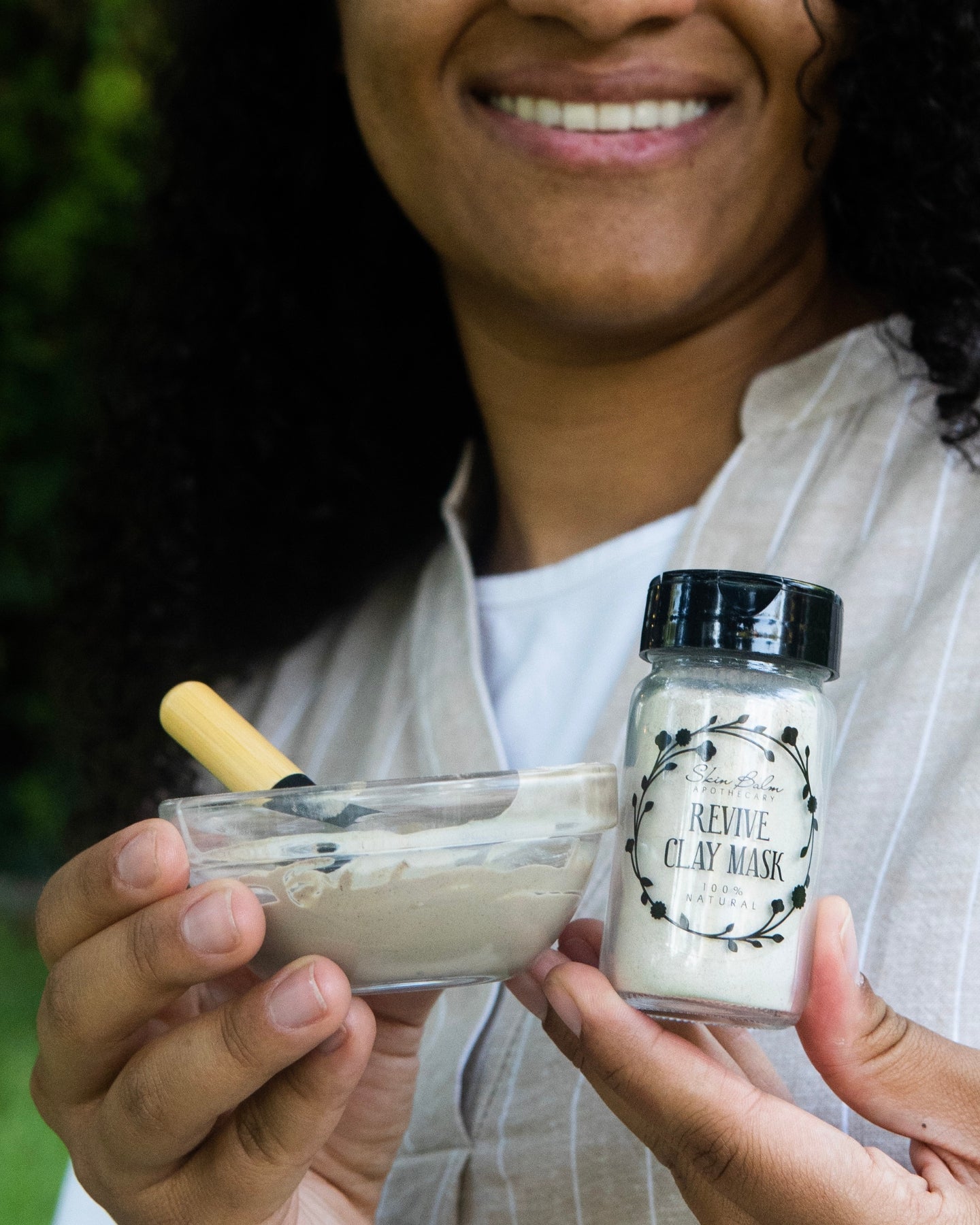 A smiling woman holds a jar of the Revive Clay Mask and a bowl of mixed product.
