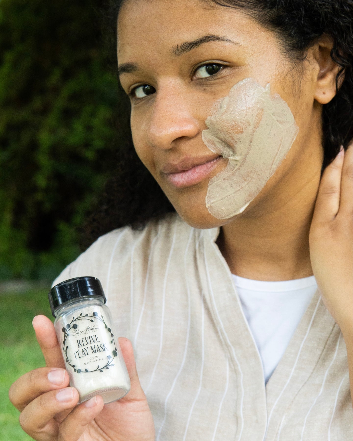 A woman with Revive Clay Mask on her face poses with a jar of the product.