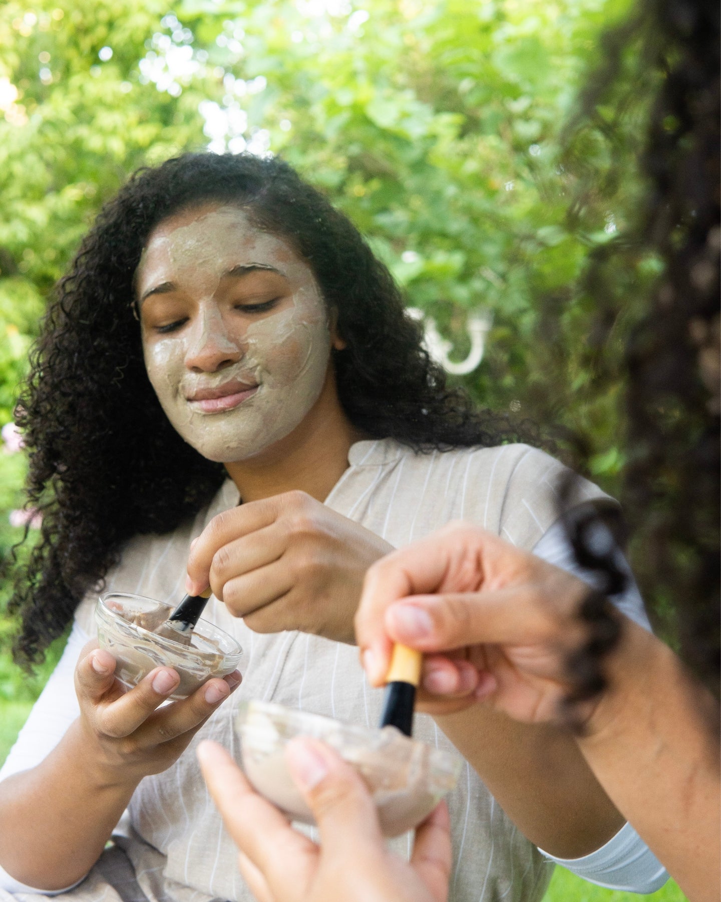 A woman with Revive Clay Mask on her face scoops mixed product out of a glass bowl.