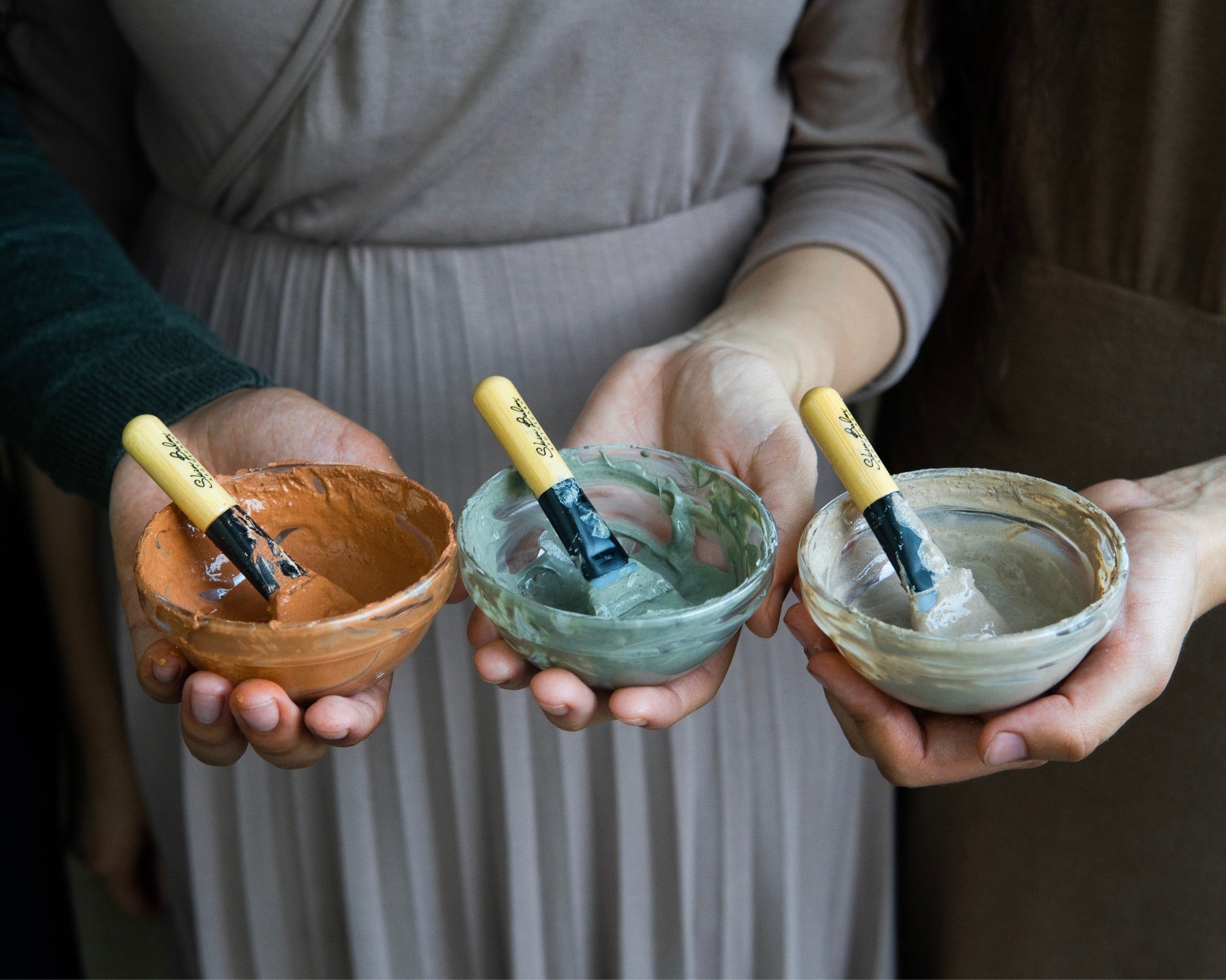 A close-up shot of three women each holding out bowls of mixed clay mask product with Silicone Applicator Brushes inside.