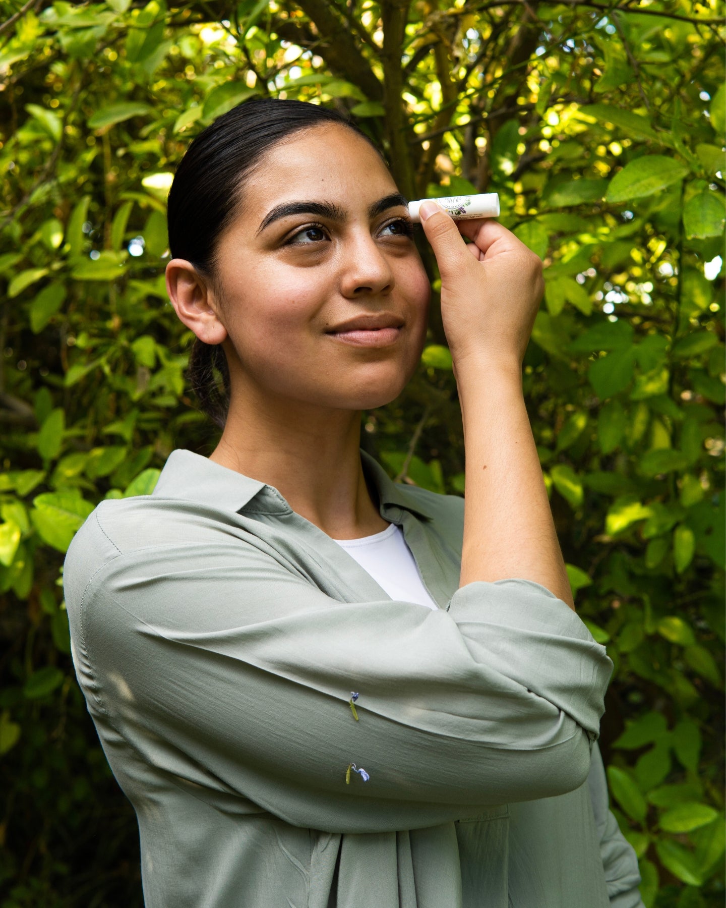A woman in a sage green dress applies The Eye Balm™ to her eyebrows.