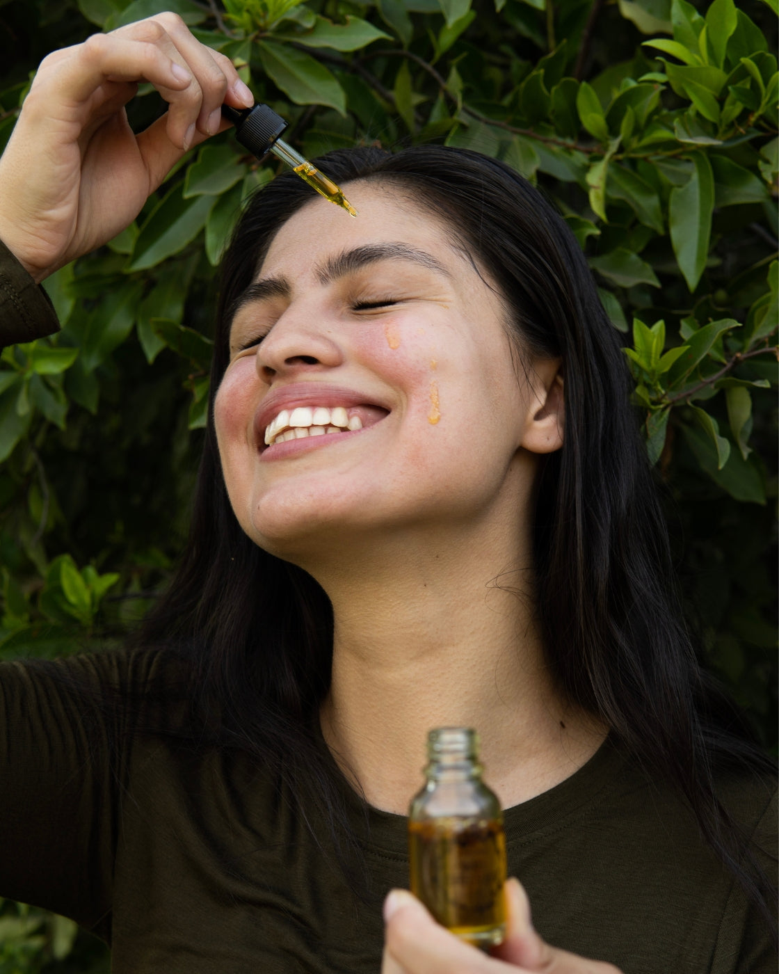 A smiling woman holds the Awake Coffee Serum with a drop of serum running down her cheek.