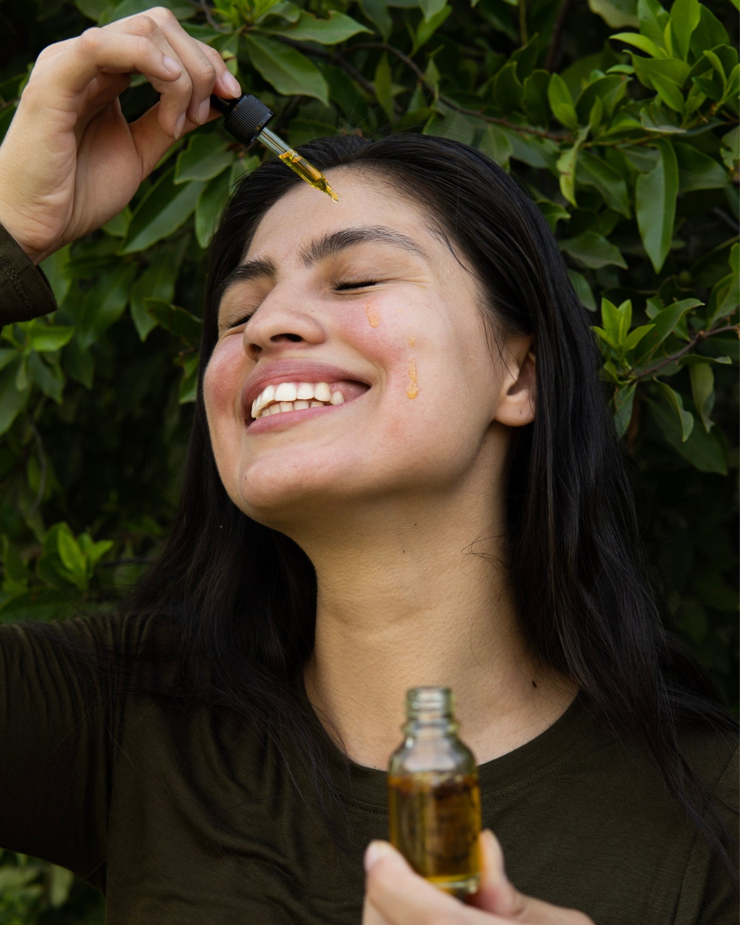 A smiling woman holds the Awake Coffee Serum with a drop of serum running down her cheek.