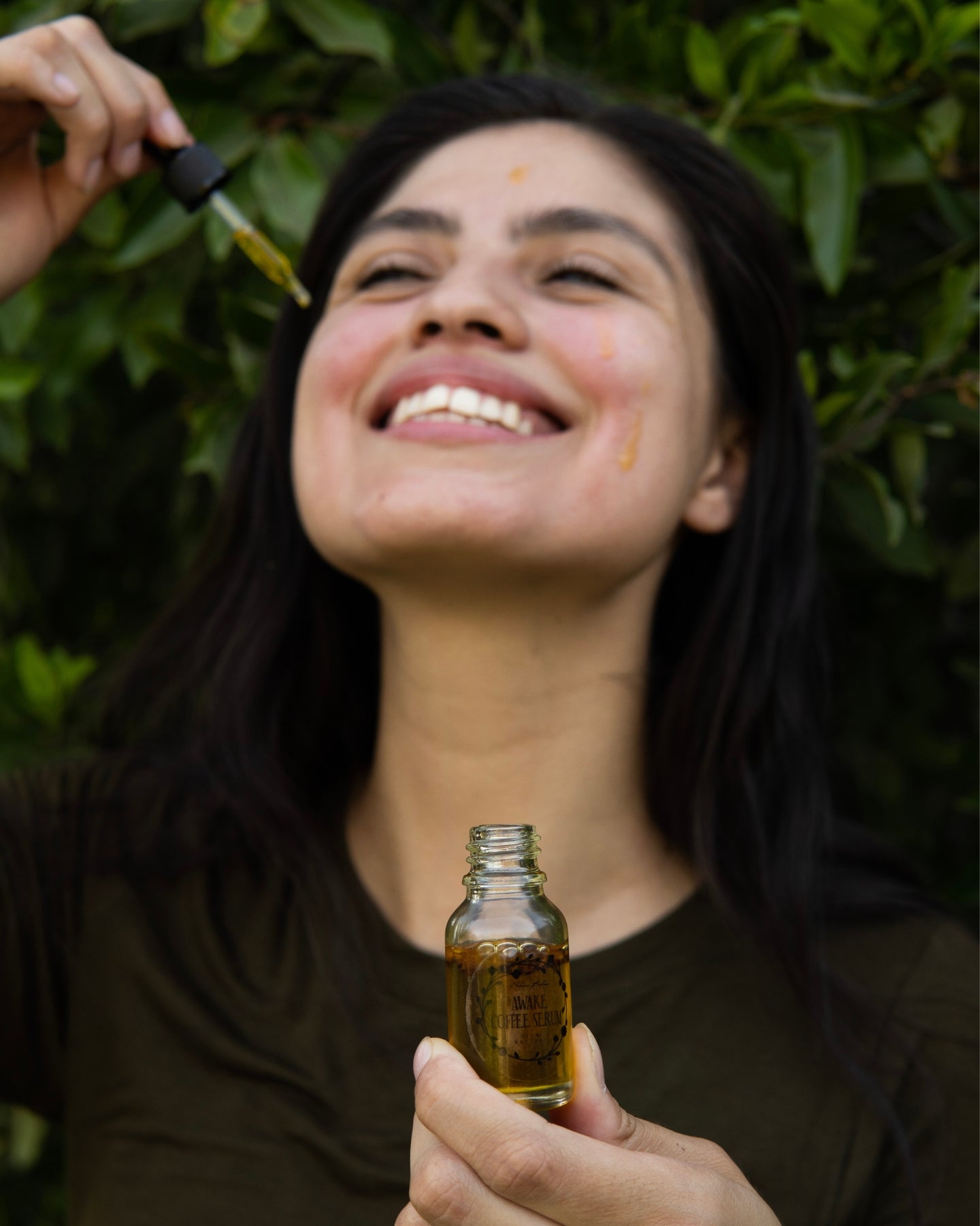 Awake Coffee Serum focused in the foreground with a smiling woman with serum on her face blurred in the background.