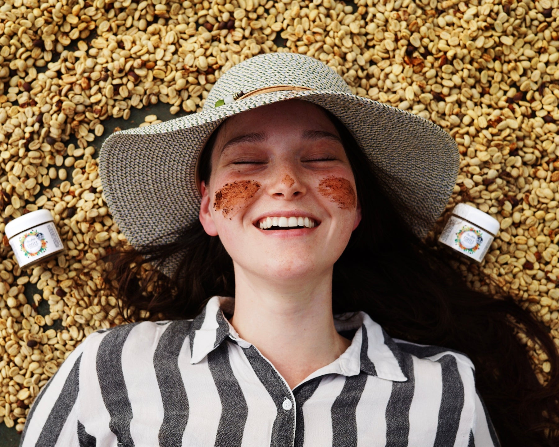 A close-up shot of a smiling woman with Coffee Face Polish on her cheeks lying down on green coffee beans.
