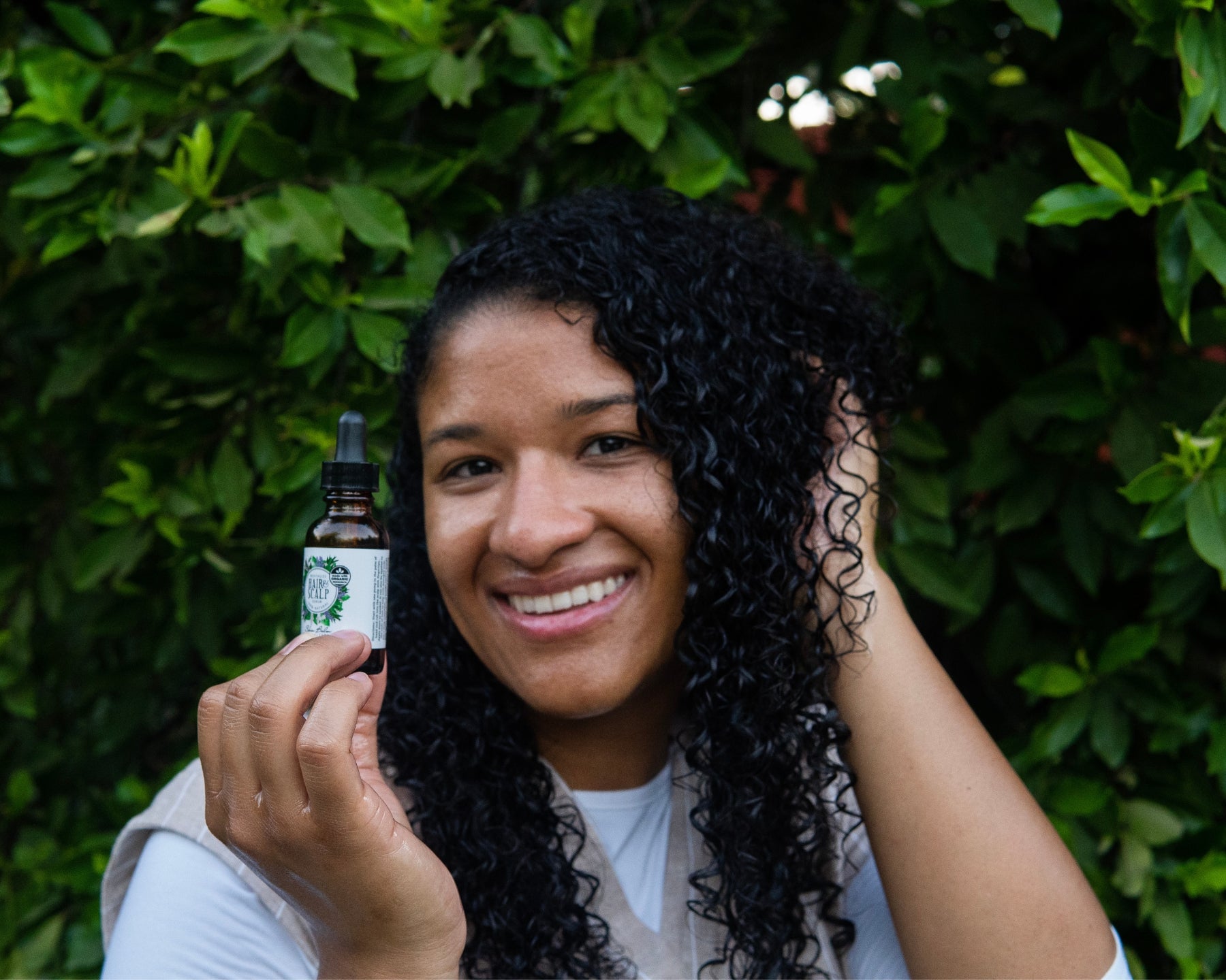 A smiling woman scrunches her hair while holding a bottle of the Revitalize Hair & Scalp Serum.