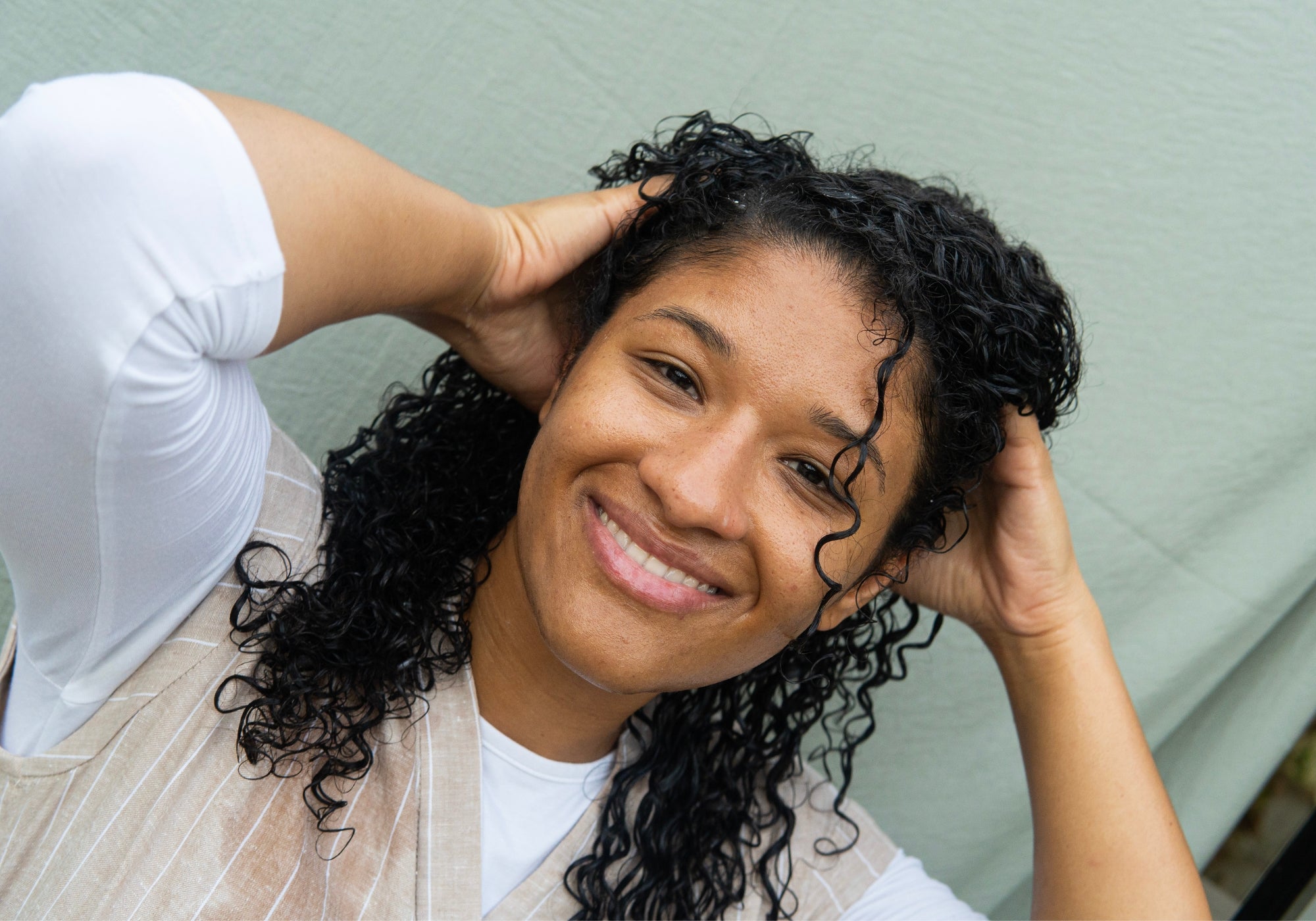 A smiling woman with Revitalize Hair & Scalp Serum in her hair scrunches her hair.